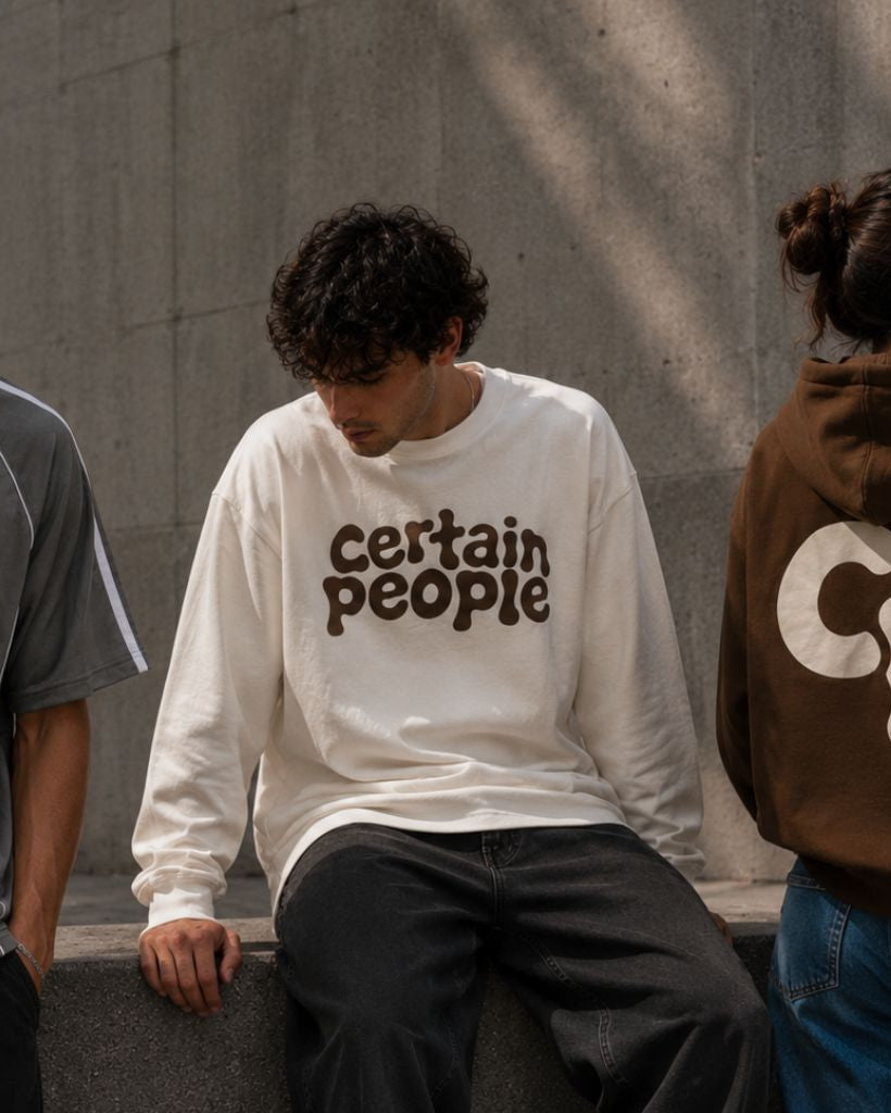 Three models wearing streetwear apparel from the brand Certain People, standing against a concrete wall. The model on the left wears a gray v-neck athletic shirt with white accents; the center model wears a white long-sleeve crewneck sweatshirt with 'certain people' printed in brown across the chest; the model on the right wears a brown hoodie showing a large white 'CP' logo on the back.