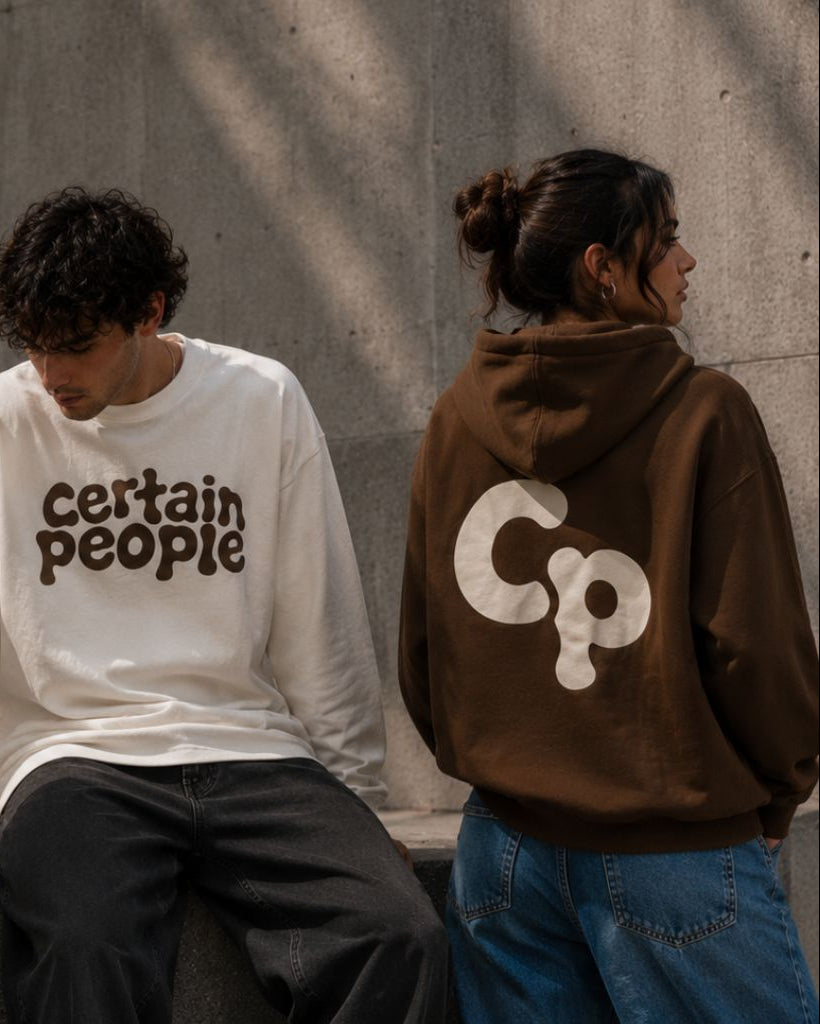 Three models wearing streetwear apparel from the brand Certain People, standing against a concrete wall. The model on the left wears a gray v-neck athletic shirt with white accents; the center model wears a white long-sleeve crewneck sweatshirt with 'certain people' printed in brown across the chest; the model on the right wears a brown hoodie showing a large white 'CP' logo on the back.