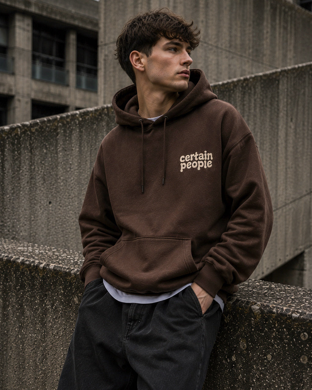 A man wearing a dark brown pullover hoodie with 'certain people' printed in beige on the left chest, leaning against a concrete wall in an urban setting.