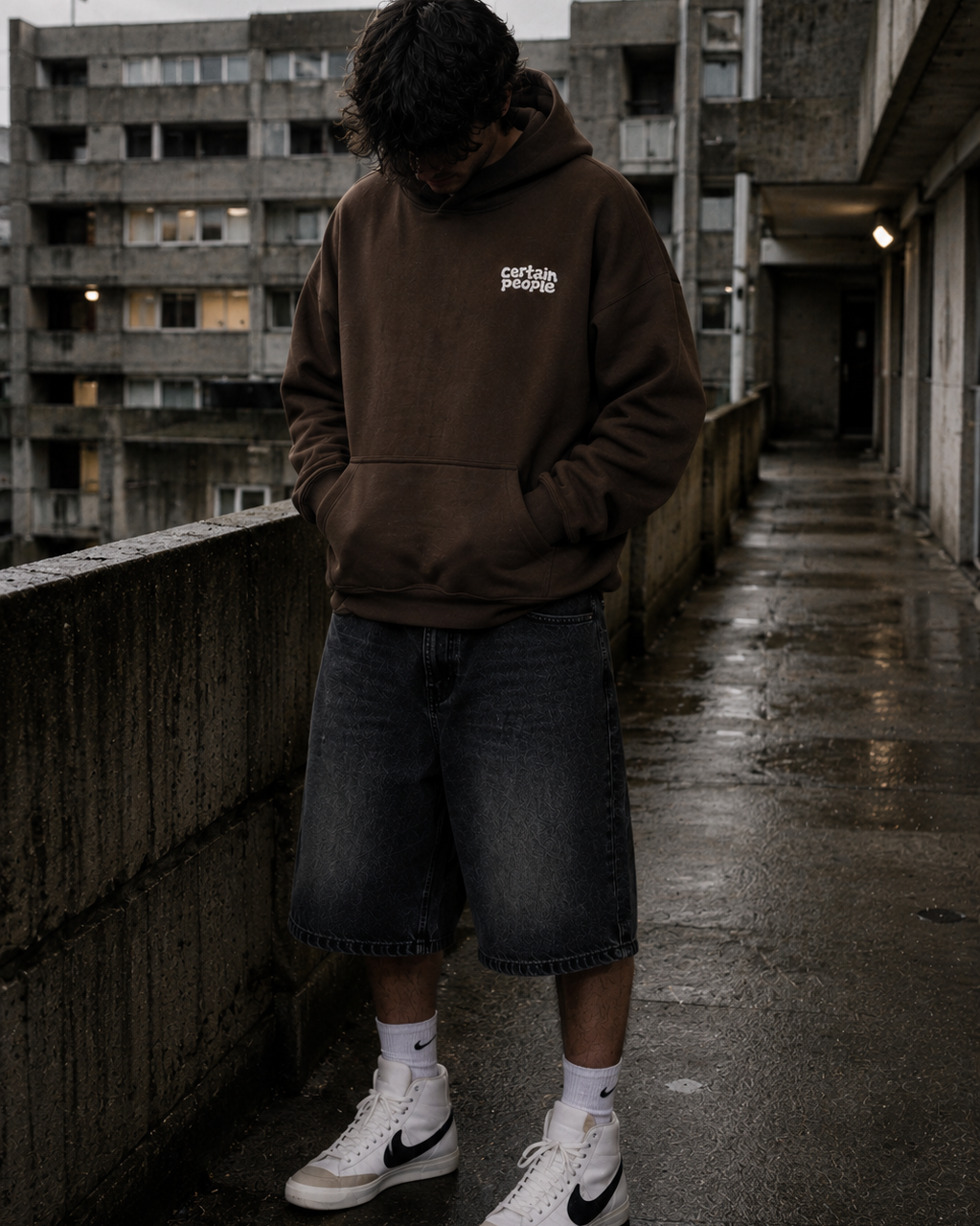 A person standing on a wet urban rooftop walkway wearing a brown pullover hoodie with a small white chest logo, dark denim shorts, white socks, and white low-top sneakers with black logos.